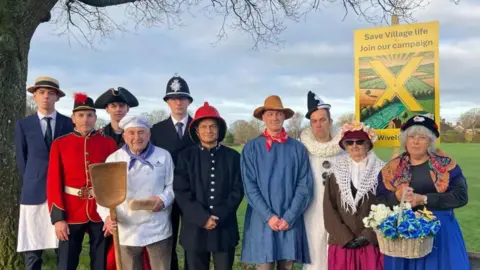 A group of people in costume in a field with a banner protesting about saving their village