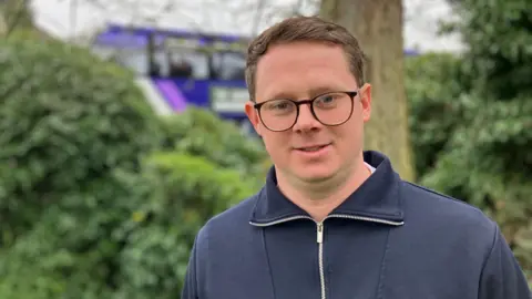 Mitchell Jones is wearing a navy zip-up jumper outdoors. He has short brown hair and glasses with dark rims. Behind him are bushes and a tree, as well as a purple bus driving by.