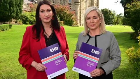 Pacemaker Emma Little-Pengelly and Michelle O'Neill holding up copies of the strategy. The backdrop is Stormont Castle and the pair are standing on the lawn. Emma Little-Pengelly is wearing a red blazer, while Michelle O'Neill is wearing a black and grey outfit