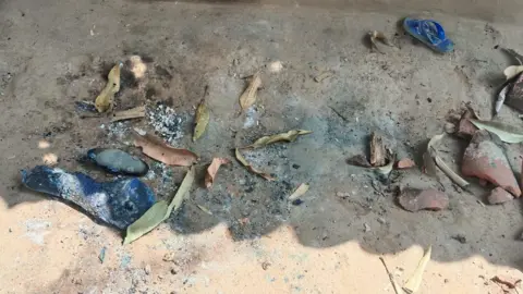 A mud floor seen with ashes, dried leaves and burnt rubber slippers