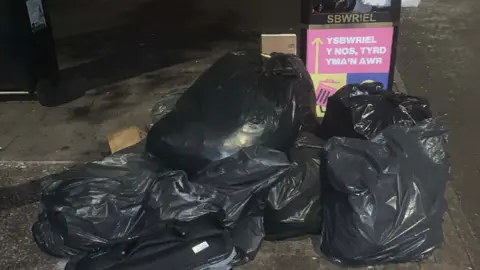 Darren Lloyd Several black bin bags dumped next to a public street bin that is full.