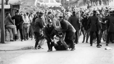 Getty Images Police and miners clash at Orgreave Colliery. In the photo a man can be seen being dragged away by two police officers.