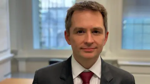 Mark Sewards, dressed in a dark suit and red tie, looks into the camera.