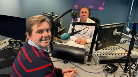 Sue Dougan/BBC Nik Johnson and Chris Mann are in the radio studio, looking at the camera, sitting opposite each other.