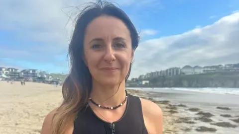 Sian has long brown hair and is wearing a black swimsuit and a necklace. She is standing on the beach at Perranporth with a blue sky above the sea and sand