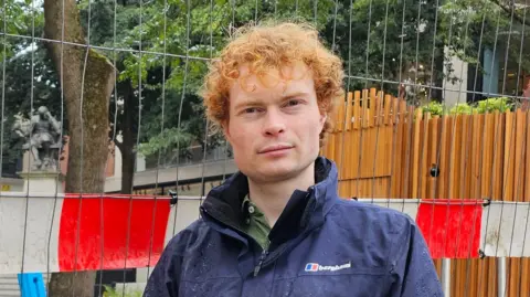 BBC Jamie Osborn is looking at the camera. On a wet day, he's wearing a blue waterproof coat. He's standing in front of fencing and construction work in Norwich city centre.