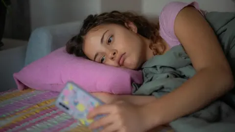 A stock image shows a young woman lying down with a glum expression as she scrolls on her smartphone
