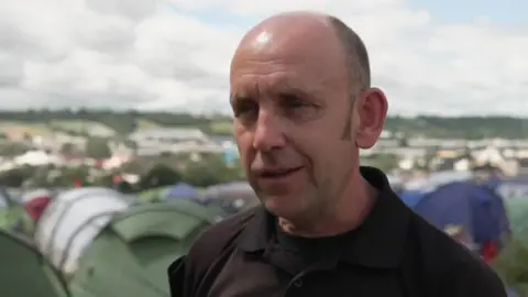Group commander Dan Searle looking to camera. He is wearing a black t-shirt and stood in front of a field of tents at Glastonbury festival.