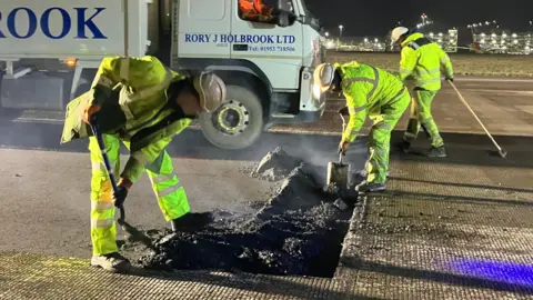 BBC/SAM READ The picture shows a well-lit airport runway at night with two construction workers in yellow high-visibility clothing digging asphalt with shovels. A third worker walks behind them. In the background a large construction lorry can be seen and in the distance a multi-storey car park.