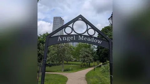 Canon Grace Thomas An iron sign at the entrance to Angel Meadow in Manchester. It is a green space with trees and apartment blocks in the distance.