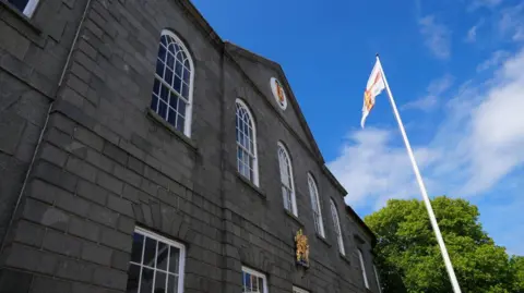 Guernsey States Chamber building St Peter Port. The building has large white oval windows and the exterior of the building is made from grey brick. There is a flag pole with the flag of Guernsey on it. The flag is white with a red cross and a gold cross within the red one.