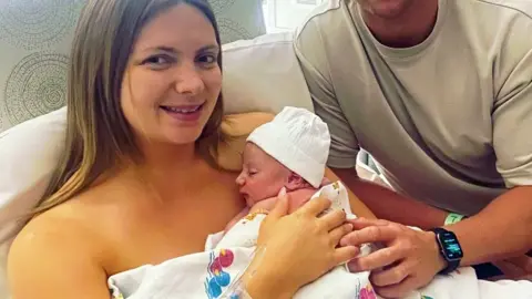 A mother with sleepig baby moments after birth. The baby has a white hat and is lying on the mothers chest