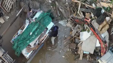 Environment Agency Two people standing in a large brown puddle and looking at a packed skip. They are surrounded by a large pile of waste.