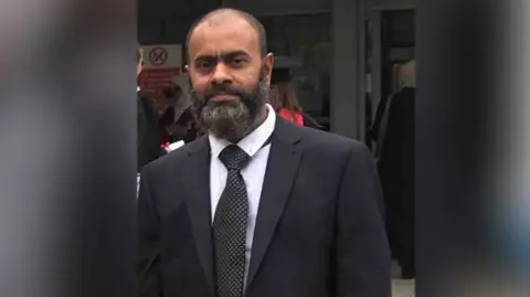 Humayra Ali Man wearing suit, has short hair, balding, with grey/black beard. Wears a black suit with tie and white shirt