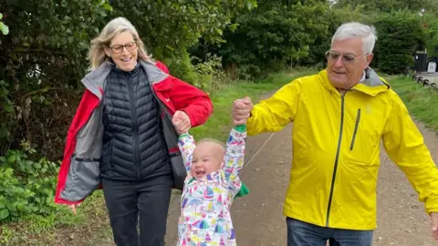 Supplied A man and a woman holding a child, with the hands of a child in the air. They are walking on a patch with bushes and grass either side of it.