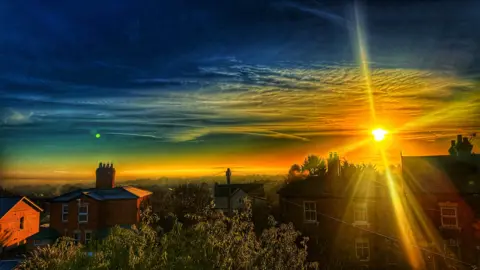 An image of a sunrise over houses in Great Malvern, with colours including green, blue and orange seen in the morning sky