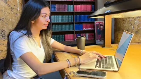 @daniistypingtoo Person seated at a wooden desk typing on a silver laptop, with a black recyclable takeaway coffee cup and a smartphone with a clear case placed either side of her. A desk lamp illuminates the workspace. In the background, shelves filled with red, green, and blue books line the wall.