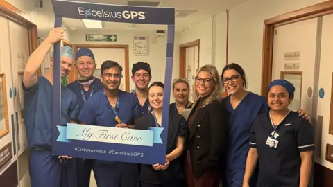NHS Somerset FT A group of smiling doctors in dark blue scrubs in a hospital corridor, holding up a large cardboard picture frame saying "my first case".