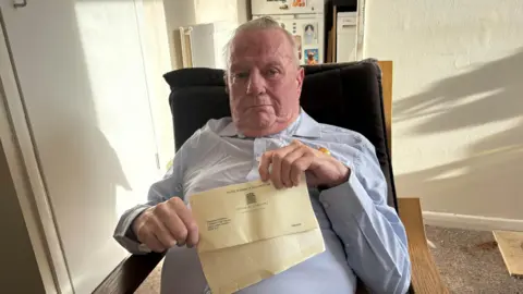 A man sat on a sofa, wearing a blue shirt. He is holding a letter which has a House of Commons logo on it.