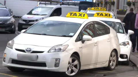 Several white taxis parked up at a rank in St Helier, Jersey. All the taxis have a yellow taxi sign on their roof.