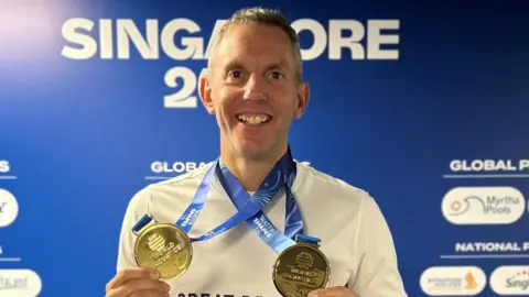 A smiling man holding two medals around his neck 