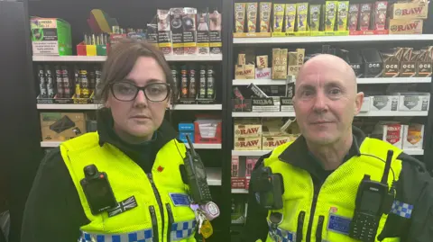 Clare Montgomery-Brown and Mark Watts in PCSO uniforms with shelves of vape products behind them