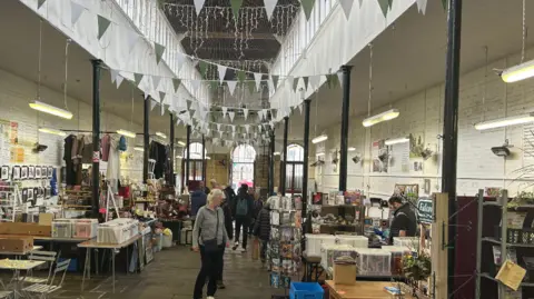 Inside the undercover market, a large room with flagstone floor, pillars down the sides, busy stalls with items and the public walking down the middle