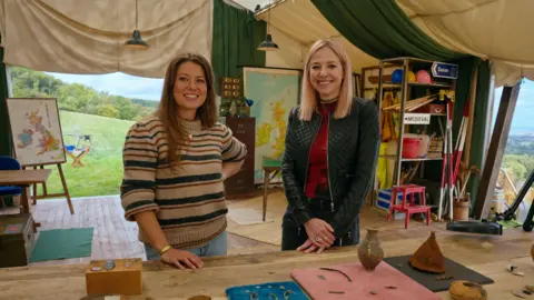 BBC/Rare TV Rosanna Price and Alice Roberts standing in the Digging for Britain tent. Rosanna is on the left, wearing a striped jumper and blue jeans and Alice on the right wearing a black leather jacket and jeans. They are standing in front of a table which has numerous small finds ranging from flints to Roman pins to pots and a clay pipe. Behind them, the tent door is open showing a field and trees and blue sky beyond. 