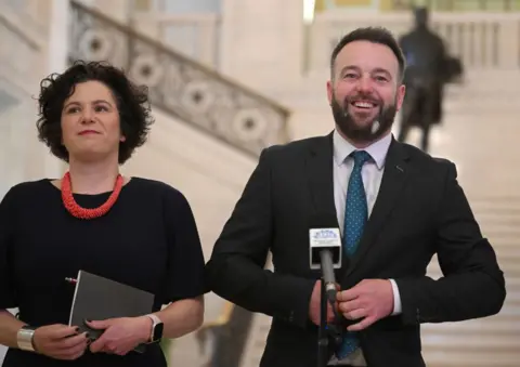 Getty Images A man stands in front of a small black microphone as he buttons up his black suit. He has short brown hair and a short brown beard, with a small white patch on it. His shirt is white and his tie in blue with small white polka dots. To his right is a woman with short brown hair. She is wearing a black dress, thick red necklace. She has a smart watch on her left hand and a gold chain on her right, she holds a small black book. Behind the pair are white stairs with a black statue of a man at the top of them. 