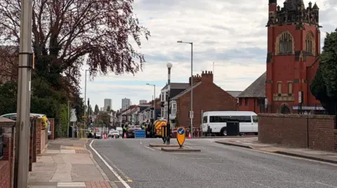Police officers speak to people in a black car just outside the cordon near the junction between Kayll Road and Chester Road in Sunderland. The cordon, which is enforced with police tape and a blue police sign, shuts off a residential street with traffic lights at the bottom of it. There is a crossing in the foreground and tower blocks can be seen in the background.