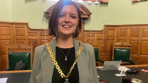 A young woman with shoulder-length brown hair. She has a nose piercing and a lip piercing. She is wearing a thick gold chain over a black and white jacket. 
