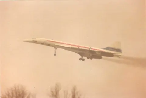 The Concorde prototype is seen taking off into the air. It is white with a red and a blue stripe.