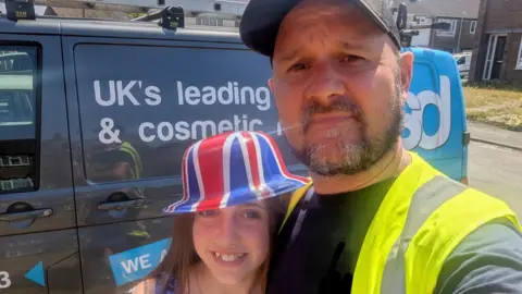 Stuart Field and his daughter take a selfie next to a grey van. He is wearing a high viz jacket and a black cap. His daughter is seen hugging him while wearing a Union Jack bowler hat. 