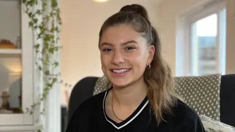 BBC Beth sits in a cushioned armchair in a living room, wearing a black sports-style top. Shelving with plants and decorative items is visible behind them, along with soft indoor lighting.