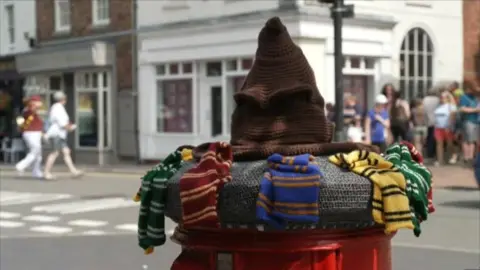 A knitted post box topper. It is a brown wizards hat, also known as the sorting hat. Around it are knitted colourful scarves of the Hogwarts houses colours - blue, red, yellow and green.