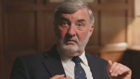 Lord Alderdice is sitting in a dimly lit room filled with wood panelling on the walls. He is wearing a dark navy, pinstripe suit with a white shirt and a house of lords tie, which is navy and contains small red and green symbols. He has grey hair swooped to the side and a grey stubbled beard. His hand is held high beside his chest as he speaks and he is wearing a gold wedding ring. 
