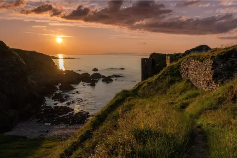Graeme Hilditch A grassy coastal path leads along a cliff overlooking a rocky cove at sunset. The sun hangs low over the sea, casting golden light across the water and surrounding hillsides. Stone walls and ruins stand on the right, while dark rocks line the shoreline below beneath a sky streaked with clouds.