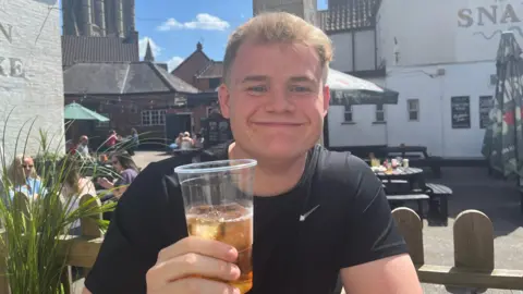 Patrycja Boryka/ BBC A man smiling and holding a drink in a plastic cup while sitting outside a pub on a sunny day. He is wearing a black t-shirt. There are people sat on tables in the background, with a cathedral behind the buildings.