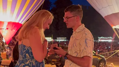 A man is proposing to a woman in front of lit up balloons.