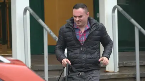 BBC Antony James is leaving a building which has steps and a handrail. His face is scrunched up and he has short dark brown hair. He is wearing a red and blue checked shirt underneath a black puffer jacket and grey jeans. He is holding a black bag. 