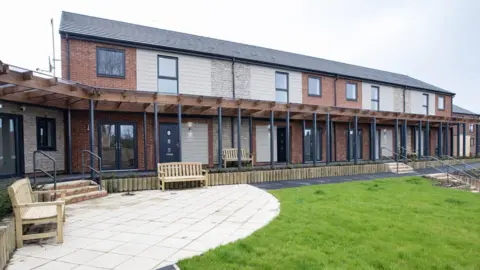 A new Gateshead Council housing development at Whitley Court, Wrekenton. It is a modern, two-storey housing block with a patio, grass and wooden benches. The units are a combination of red brick, white and grey stone. A wooden canopy structure sits between the ground and first floors, supported by dark grey pillars.