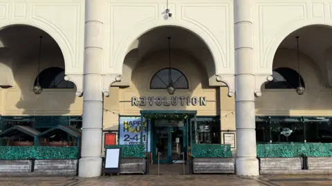 Elliot Deady/BBC A photo of the entrance to Revolution nightclub taken from the road. It is a large cream-coloured building with fake, green bushes in front of it. The Revolution logo is located above the doorway. 