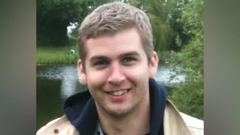 Contributed Alex Young stands in front of a pond. He has short blonde hair and wears a dark hoodie with a cream jacket over the top. 