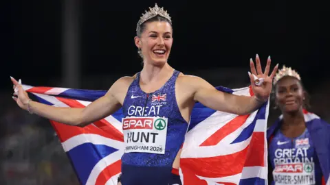 She is smiling in the middle of the photo and looking away from the camera while wearing a tiara and the national flag.  Dina-Asher Smith is on the right in the background.