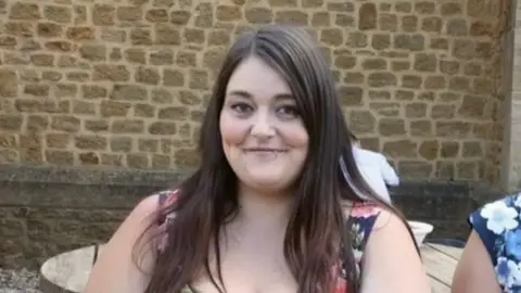 Charlotte Avis is sat smiling. She has long, brown hair and has a ring in her bottom lip.