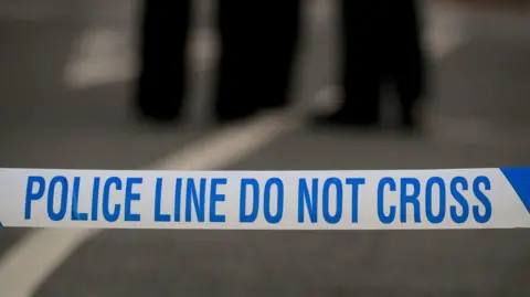 A close up of police tape that reads "police line do not cross".  The legs of two police officers are blurred in the background.