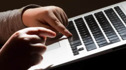 PA Media A child's hands typing on a laptop. The laptop's keyboard is black and the child is wearing a long sleeve grey top.