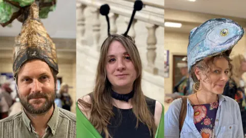 A man with a paper tree on his head on the left a woman dressed as a butterfly in the middle and a woman with a paper fish on her head on the right