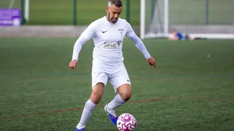 SANDS FC NOTTINGHAM Man playing football. He is wearing white football kit and blue boots.