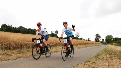 Two men cycling along a lane in a field on a charity bike ride. They are wearing blue and white lycra tops, black shorts, and red helmets. Their bikes are red and have a blue and white number card on the handlebars. The men are waving and smiling. It is a cloudy day but the grass on either side of the lane is scorched.